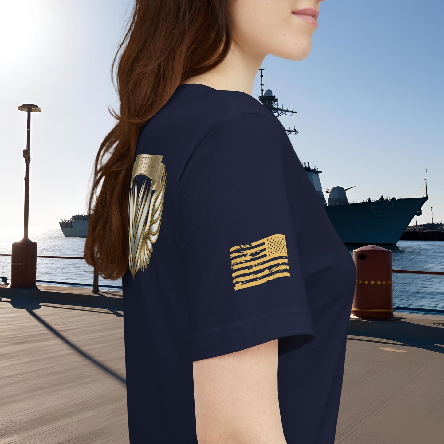 Person wearing a navy blue shirt with gold eagle and American flag design, standing on a dock with ships in the background.