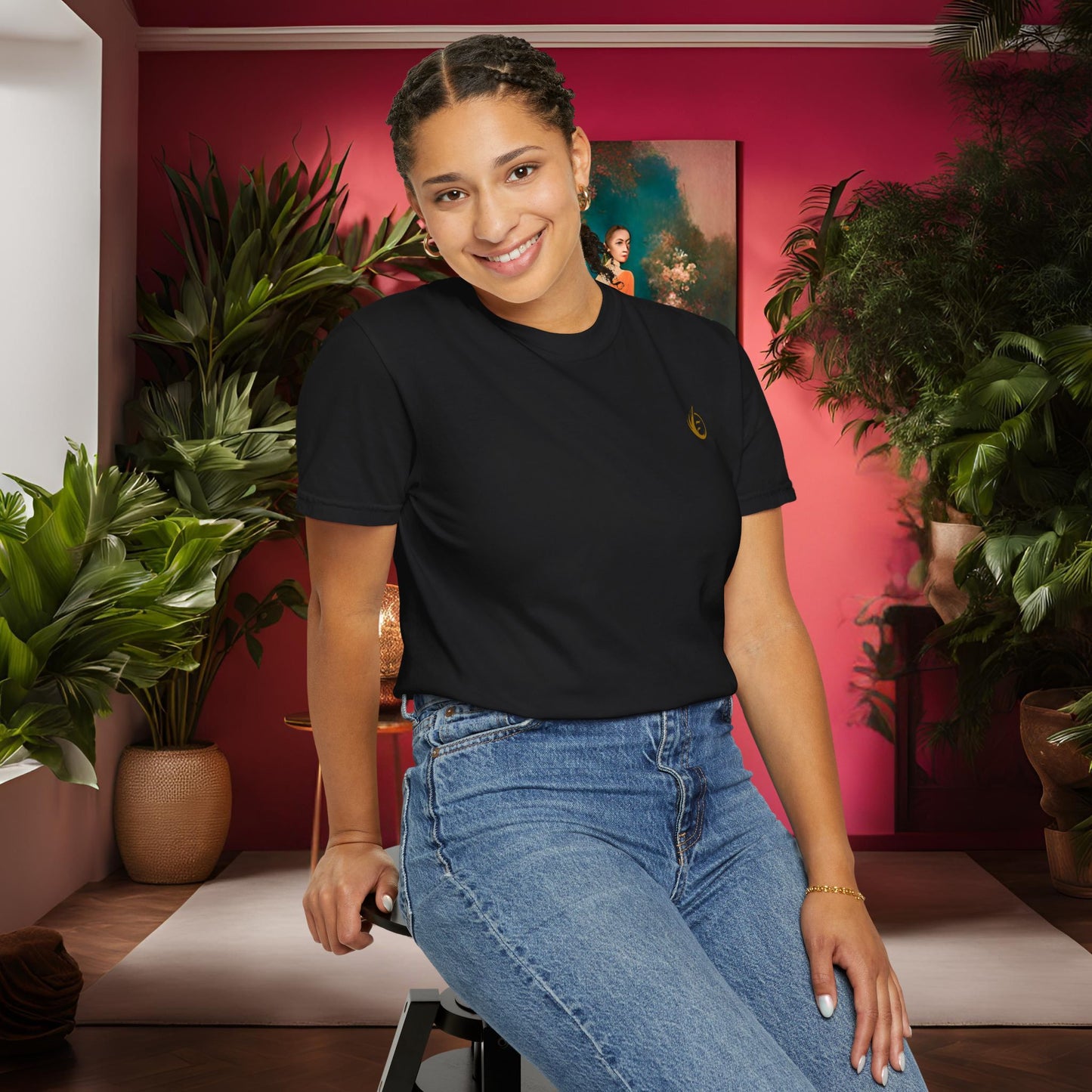 Woman in black 'EssenceInnoCreate' t-shirt and blue jeans sitting on a stool in a room with plants and a pink wall.