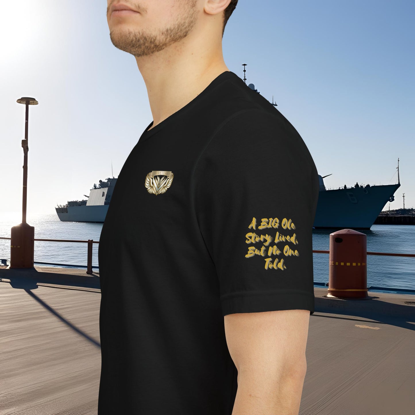 Person wearing a black t-shirt with gold text and logo by a waterfront with ships in the background