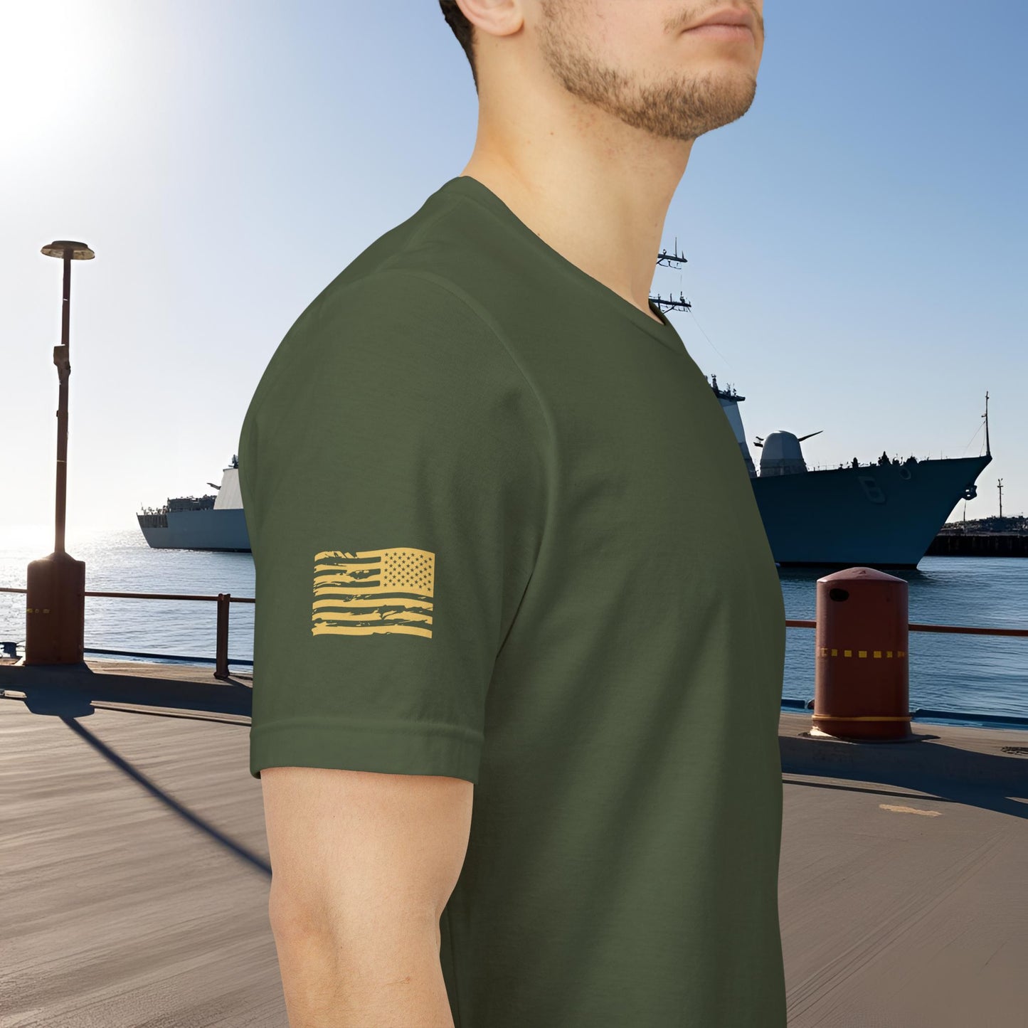 Person wearing a green t-shirt with a flag design by a waterfront with ships in the background.