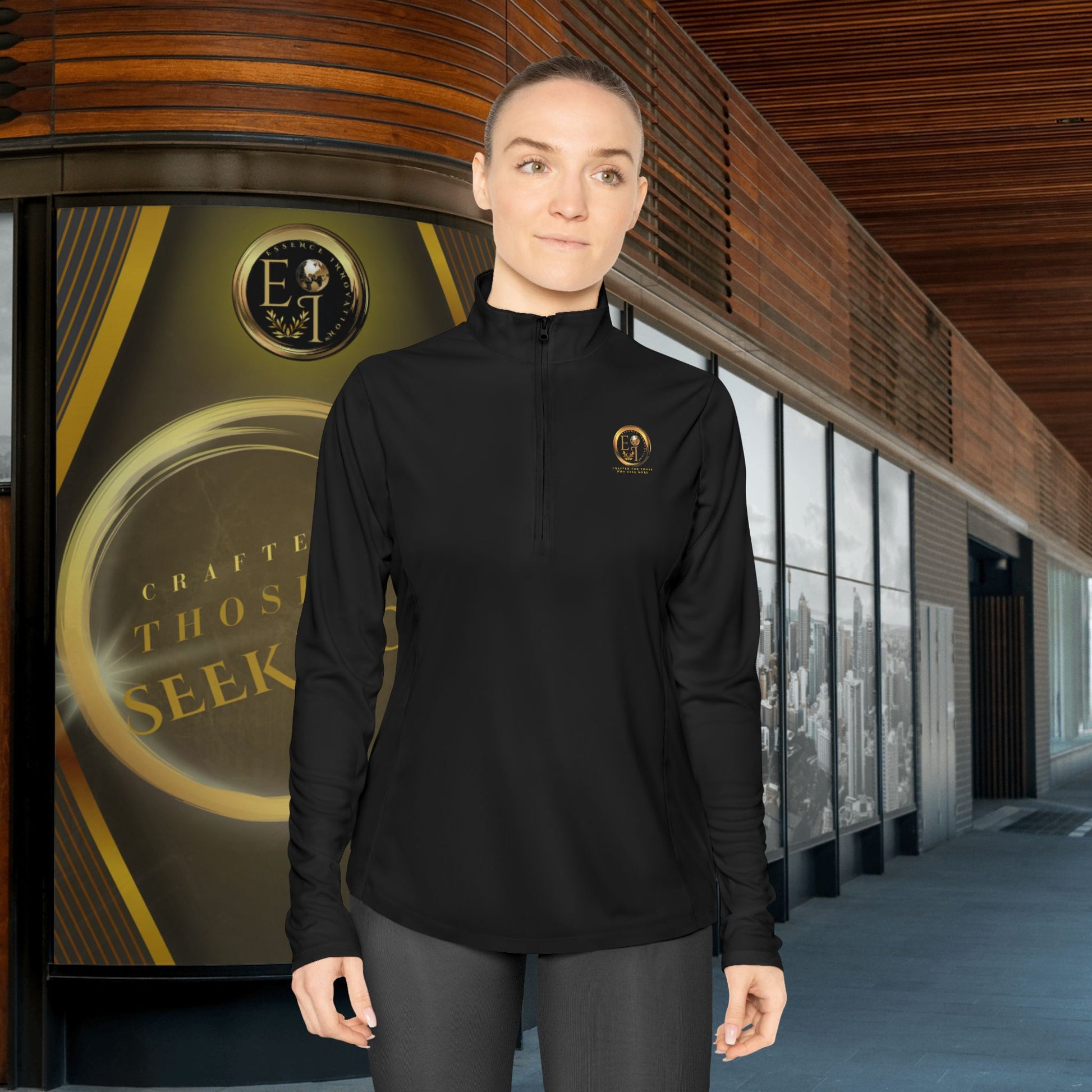 Person wearing a black Essence Innovation Seek More quarter-zip with a logo in front of a building with a large bottle-like structure.