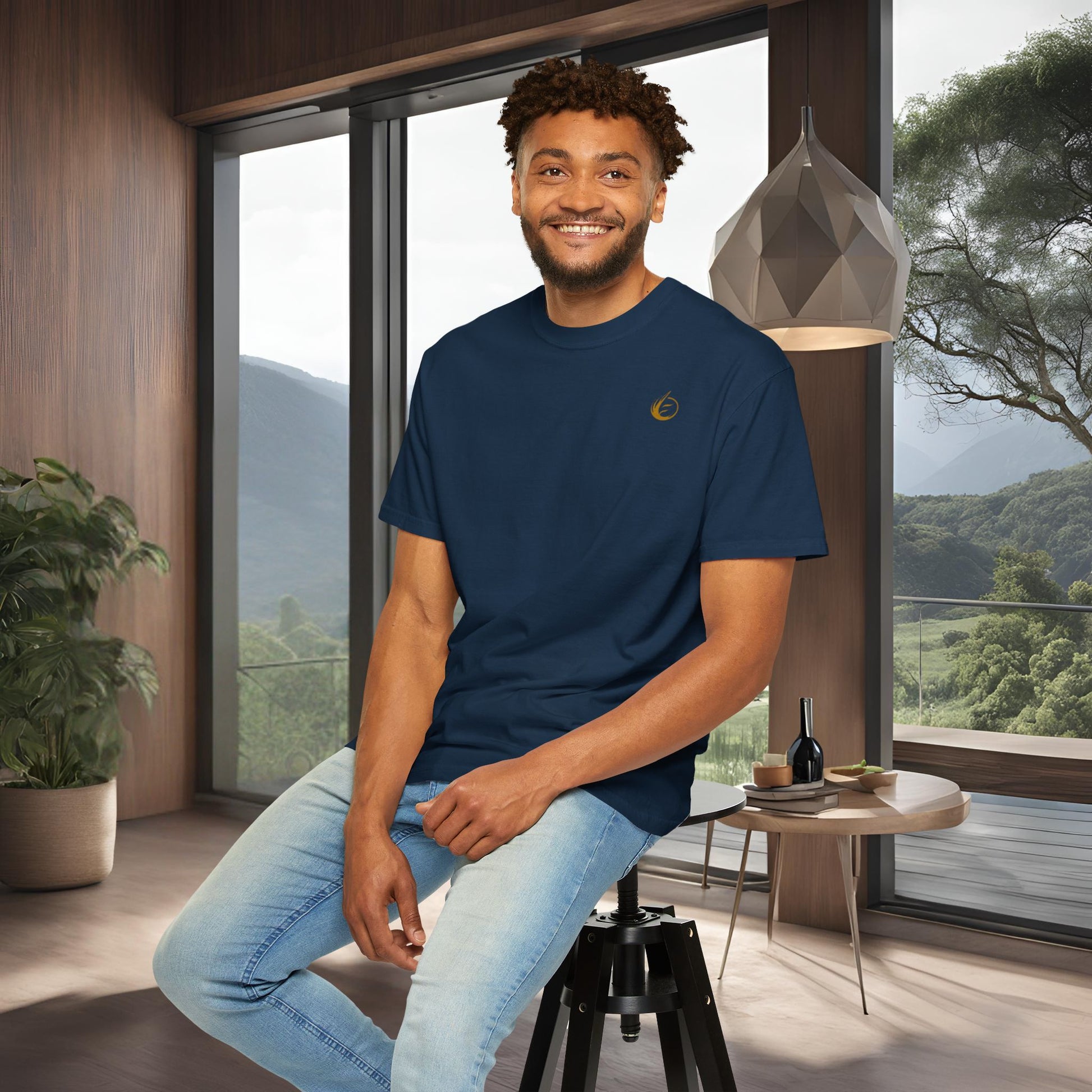 Man sitting on a stool in a modern room with large windows and a scenic view in a 'EssenceInnoCreate' T-shirt.
