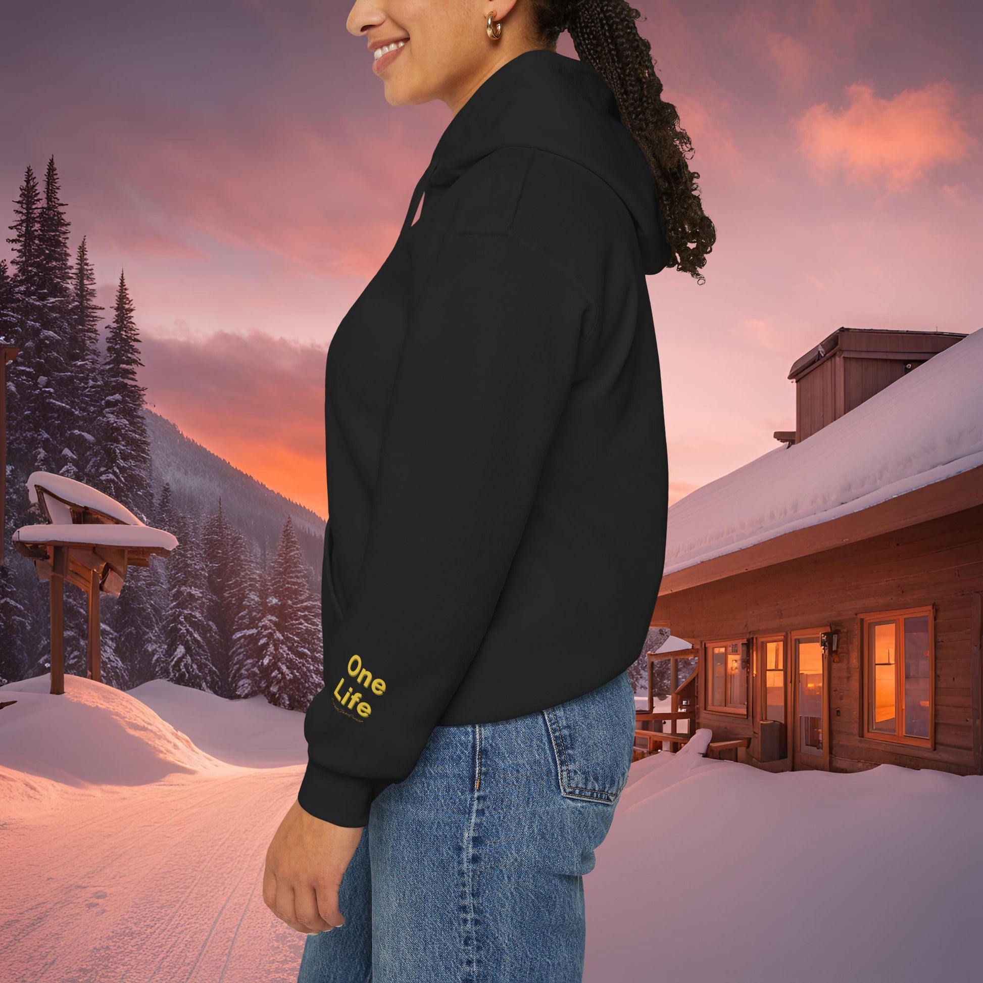 Person wearing a black Essence Innovation hoodie with 'One Life' text in a snowy landscape with a cabin and sunset.