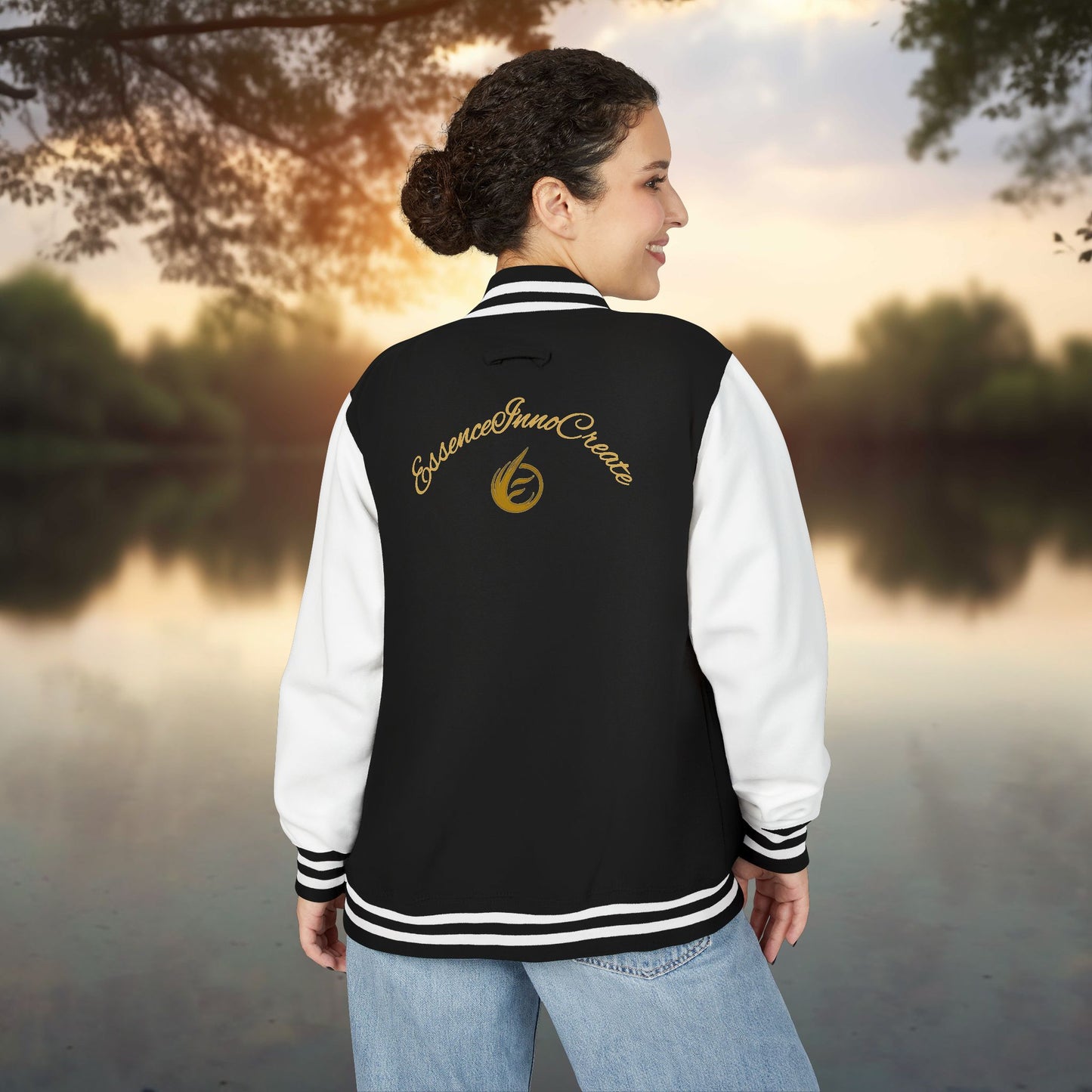 Person wearing a black and white letterman jacket with a EssenceInnoCreate logo, standing by a lake at sunset.