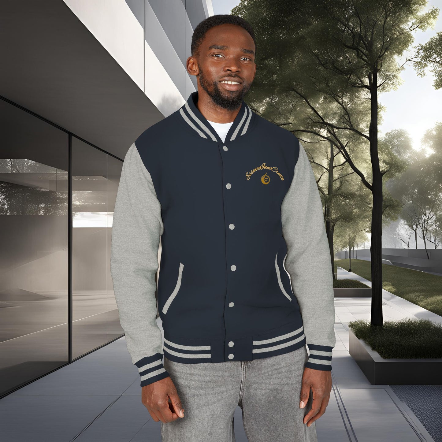 Man wearing a navy and gray varsity 'EssenceInnoCreate' jacket standing outdoors with modern building and trees in the background