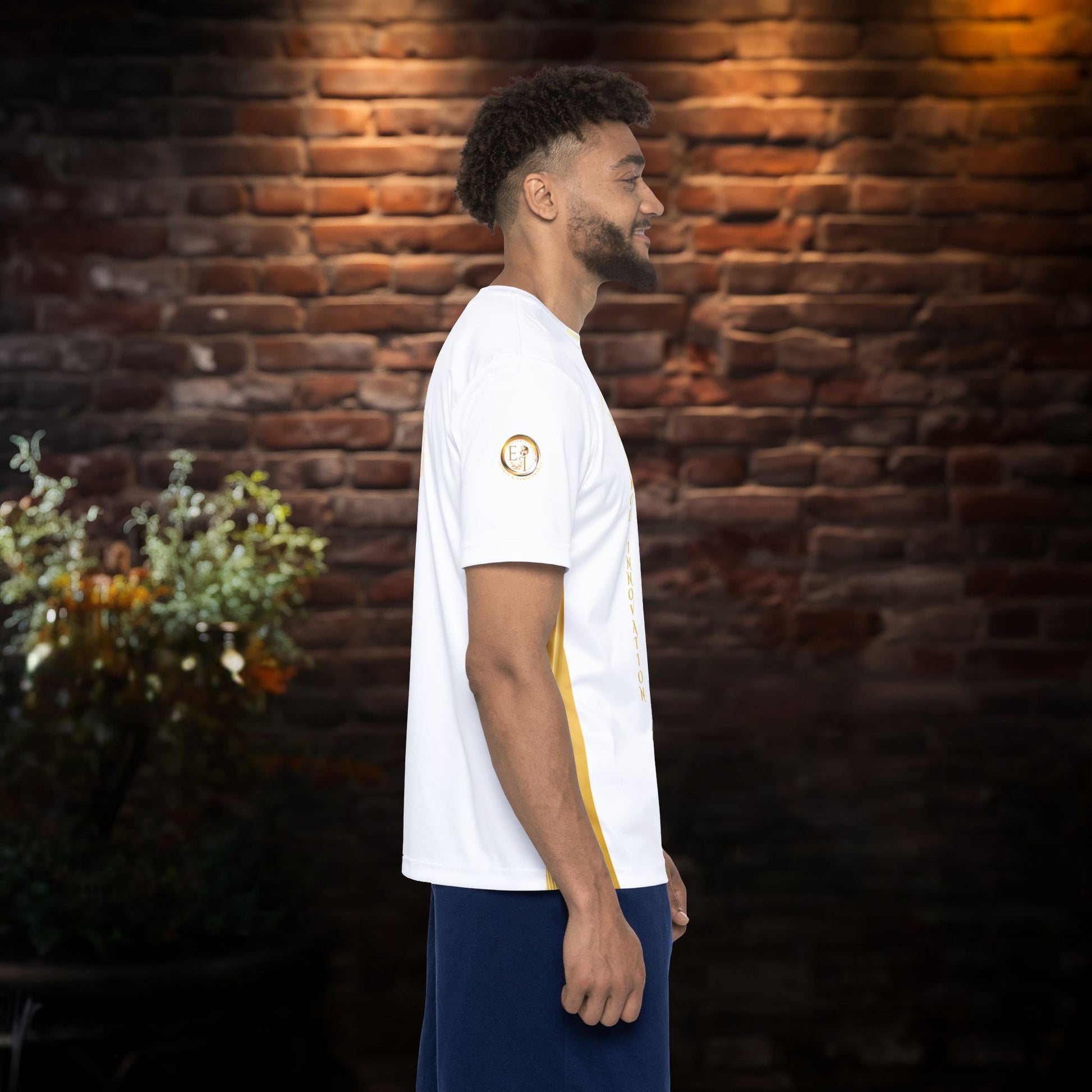 Man wearing a white t-shirt with gold Essence Innovation logo and accents against a brick wall.