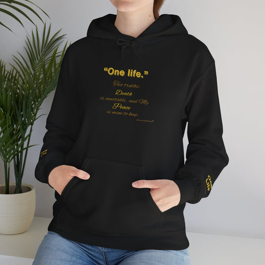 Person wearing a black Essence Innovation 'One Life' hoodie with a white cross design, standing indoors with a plant in the background.