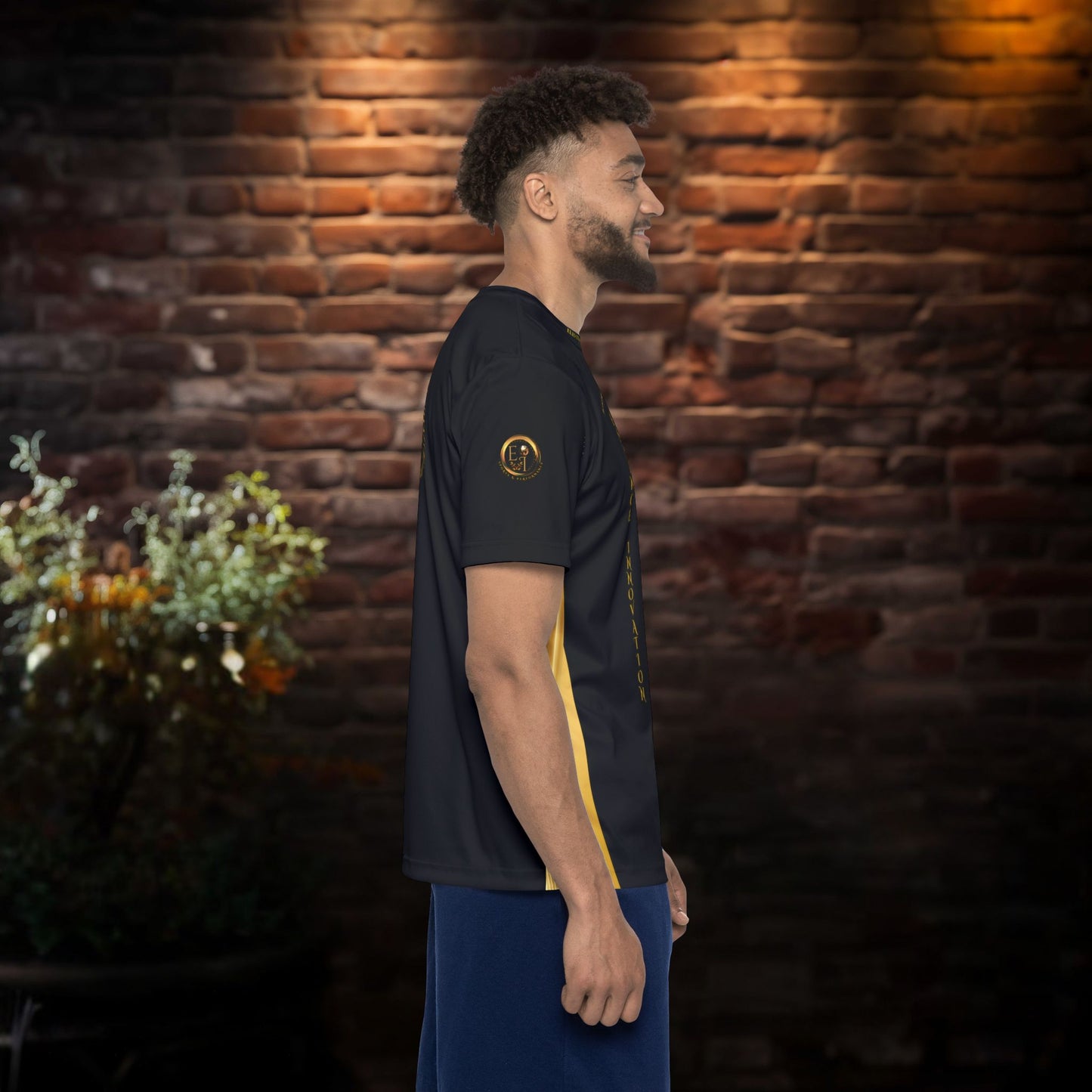 Man wearing a black t-shirt with gold Essence Innovation logo and designs against a brick wall.