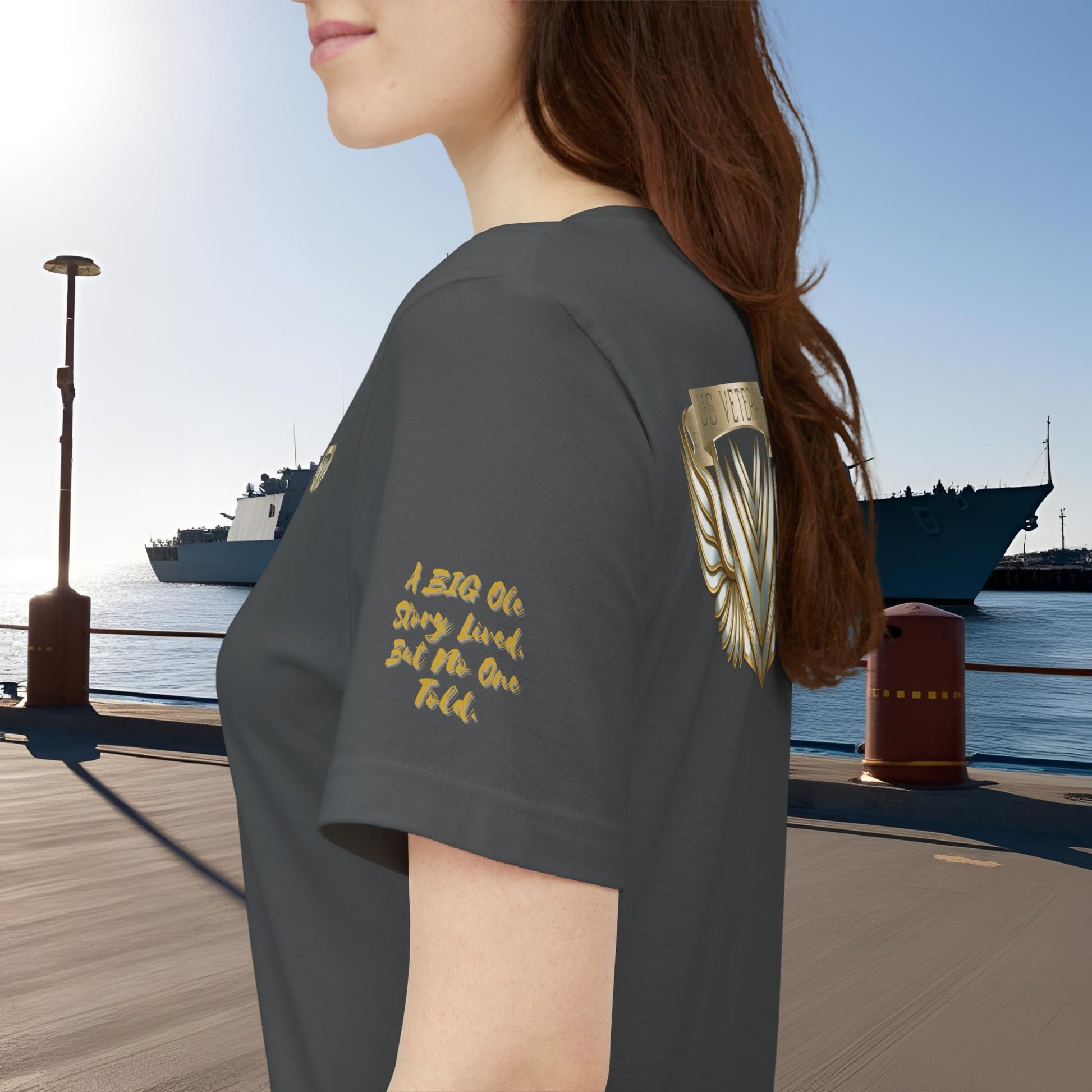 Person wearing a dark gray t-shirt with gold text and design, standing by a waterfront with ships in the background.