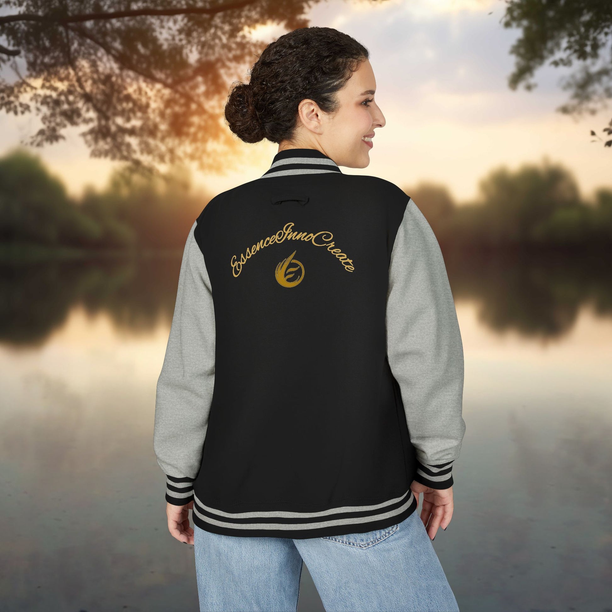 Person wearing a black and gray varsity jacket with 'EssenceInnoCreate' logo by a lake at sunset.