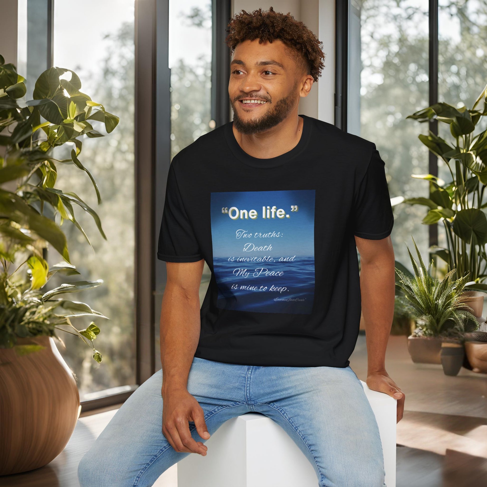 Man wearing a black 'One Life' t-shirt with a blue design, sitting in a room with plants.