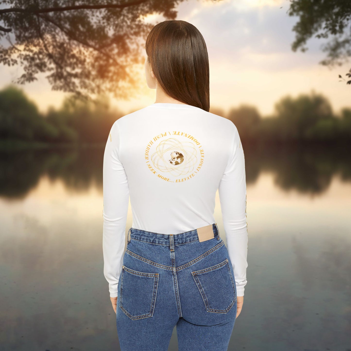 Person wearing a white long-sleeve Essence Innovation Seek More shirt with a logo and blue jeans, standing by a lake at sunset.