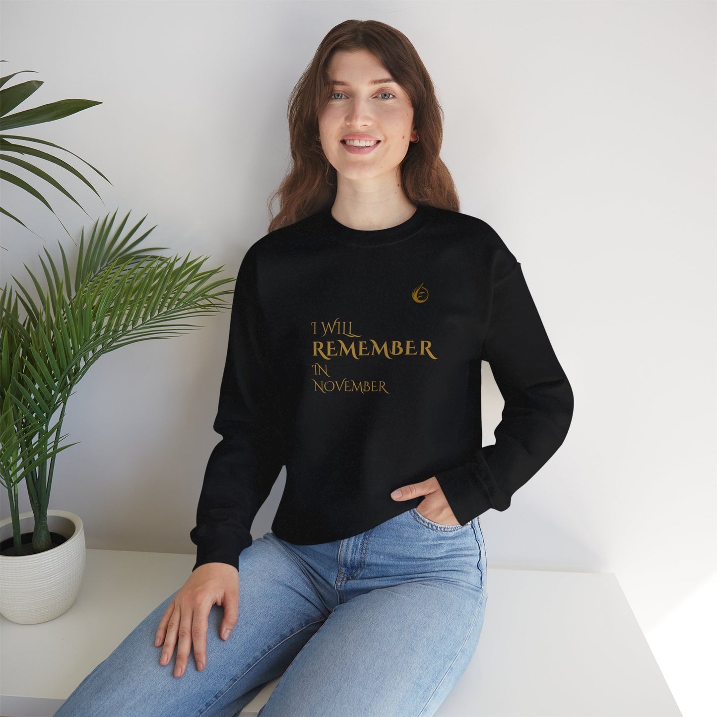 Person wearing a black sweatshirt with 'I WILL REMEMBER IN NOVEMBER' text, sitting against a white wall with a plant.