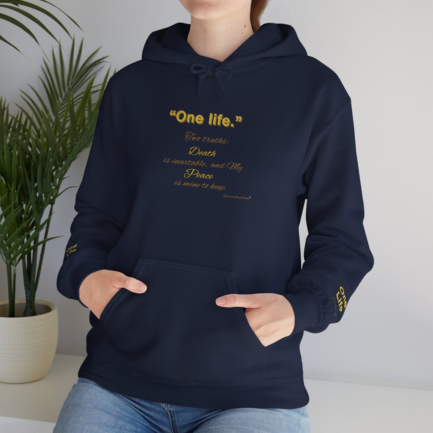 Person wearing a navy blue Essence Innovation 'One Life' hoodie with colorful graphics and text, standing indoors with a plant in the background.