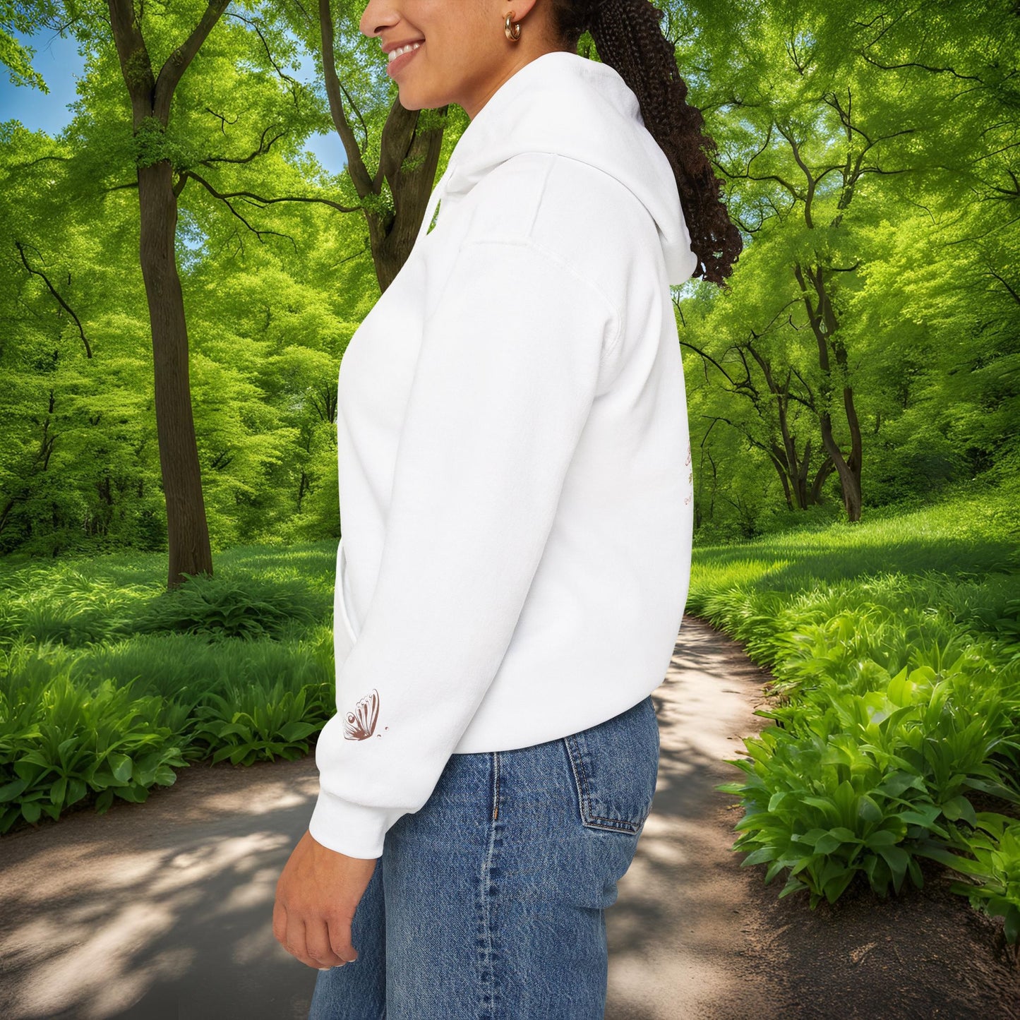 Person wearing a white Essence Innovation Bohemian Butterfly Crest hoodie and blue jeans standing in a lush green park.
