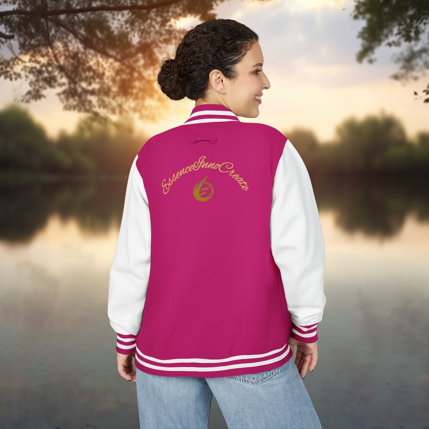 Person wearing a pink and white varsity jacket with EssenceInnoCreate text, standing by a body of water.