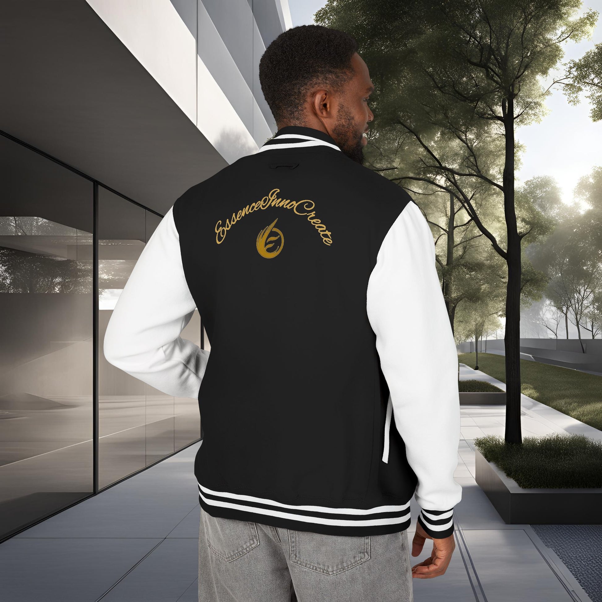 Man wearing a black letterman jacket with a EssenceInnoCreate logo, standing in an urban setting with modern architecture and trees.