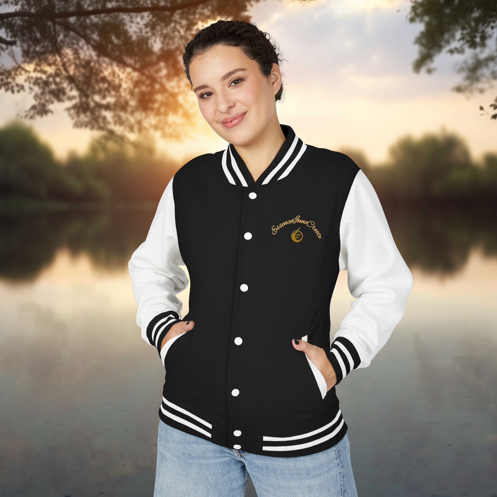 Person wearing a black and white varsity jacket with a EssenceInnoCreate logo, standing outdoors with a scenic background.