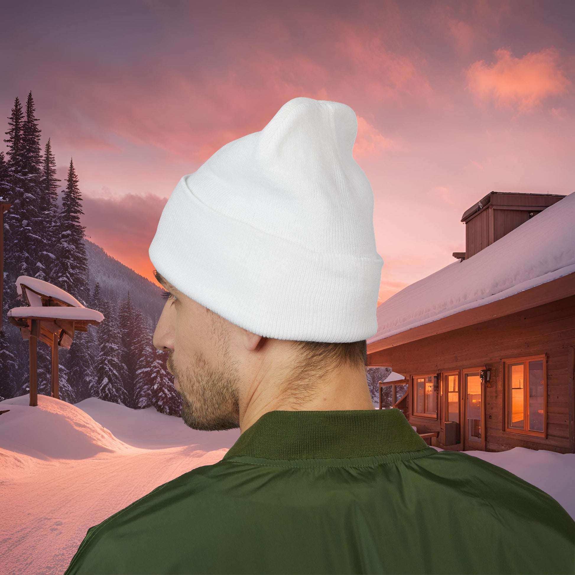 Person wearing a white EssenceInnoCreate beanie and green jacket in a snowy landscape with a wooden cabin and sunset sky.