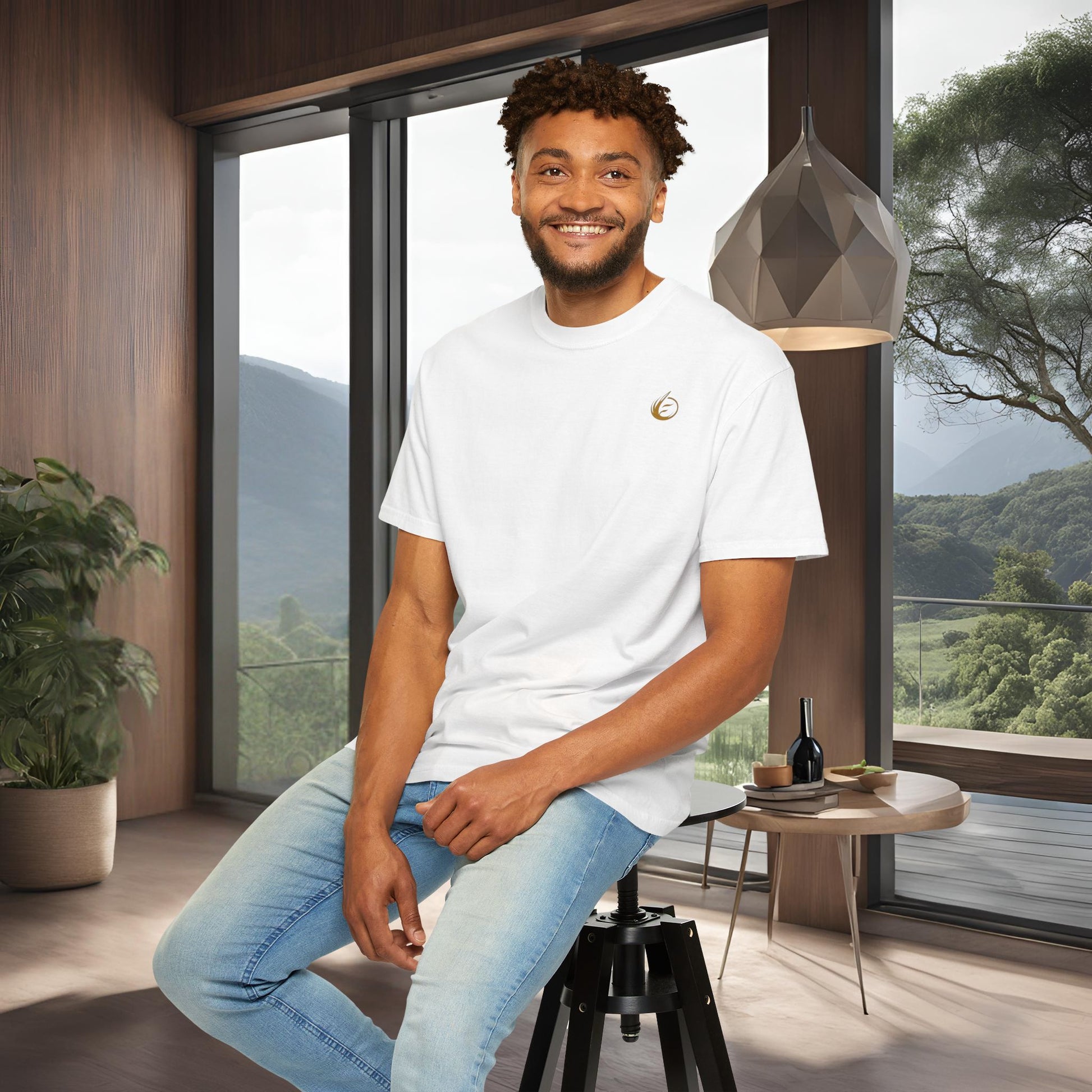 Man sitting on a stool in a modern room with large windows and a scenic view in a 'EssenceInnoCreate' T-shirt.