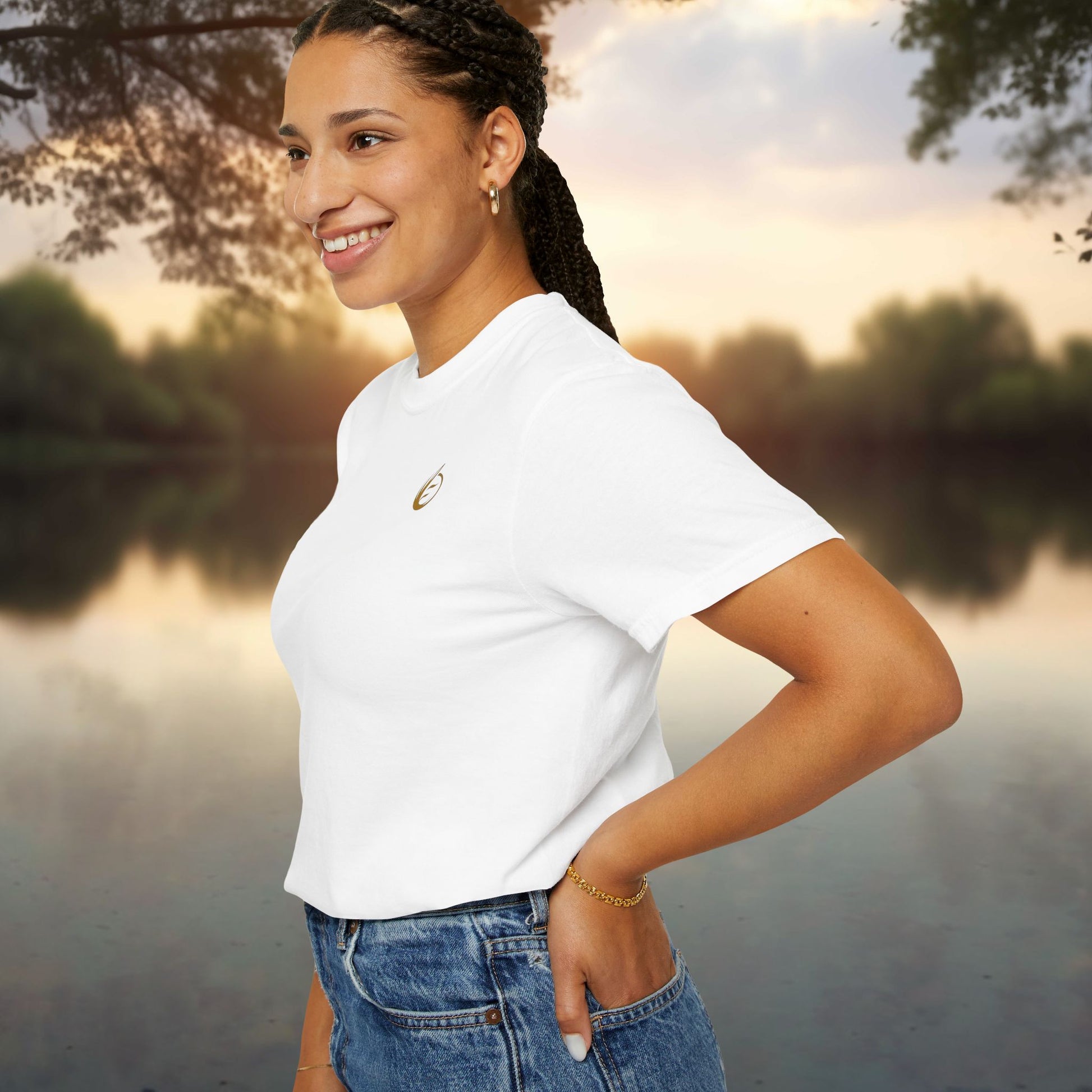 Woman in a white 'EssenceInnoCreate' t-shirt and blue jeans standing by a lake with trees in the background