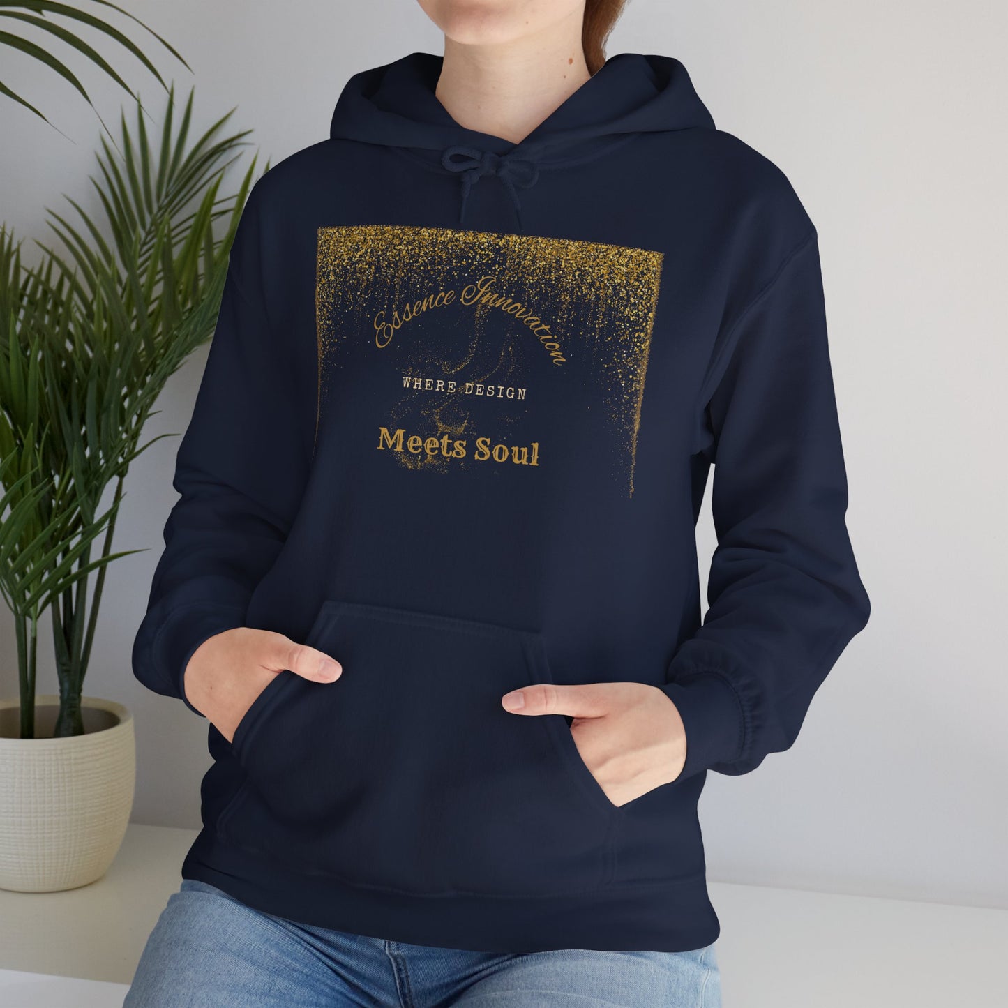 Person wearing a black hoodie with a gold Essence Innovation 'Design Meets Soul', standing indoors with a plant in the background.