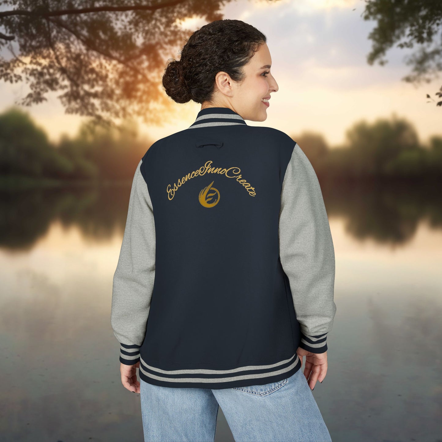 Person wearing a navy and gray varsity jacket with 'EssenceInnoCreate' logo, standing by a lake at sunset.