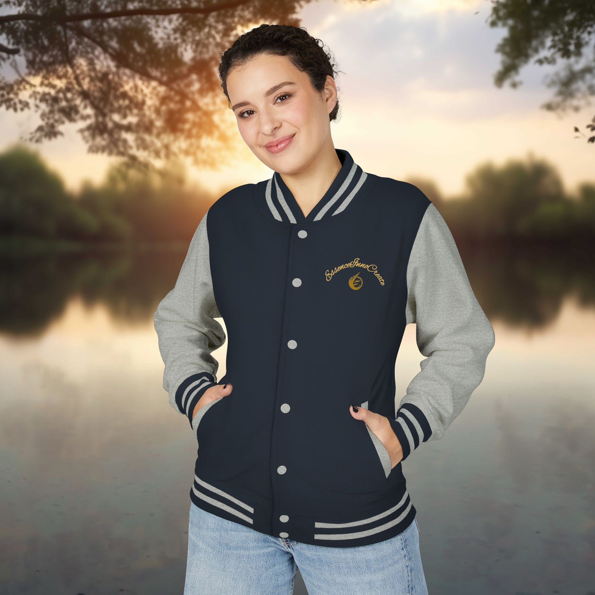 Person wearing a navy and gray varsity jacket with a 'EssenceInnoCreate' logo, standing outdoors with a scenic background.