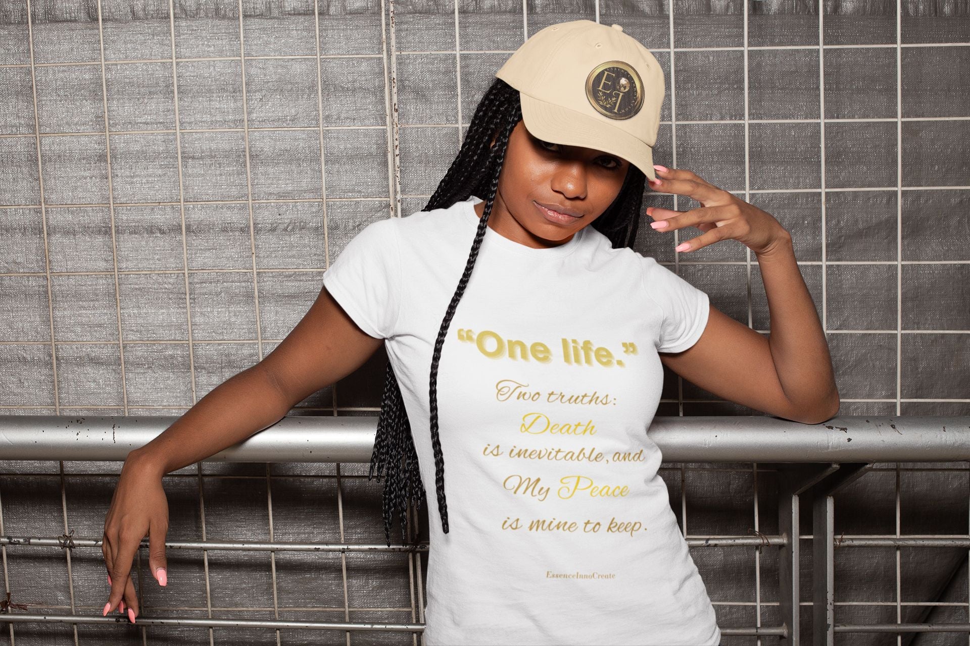 Person wearing a white One Life t-shirt with text and a beige cap in front of a tiled wall.